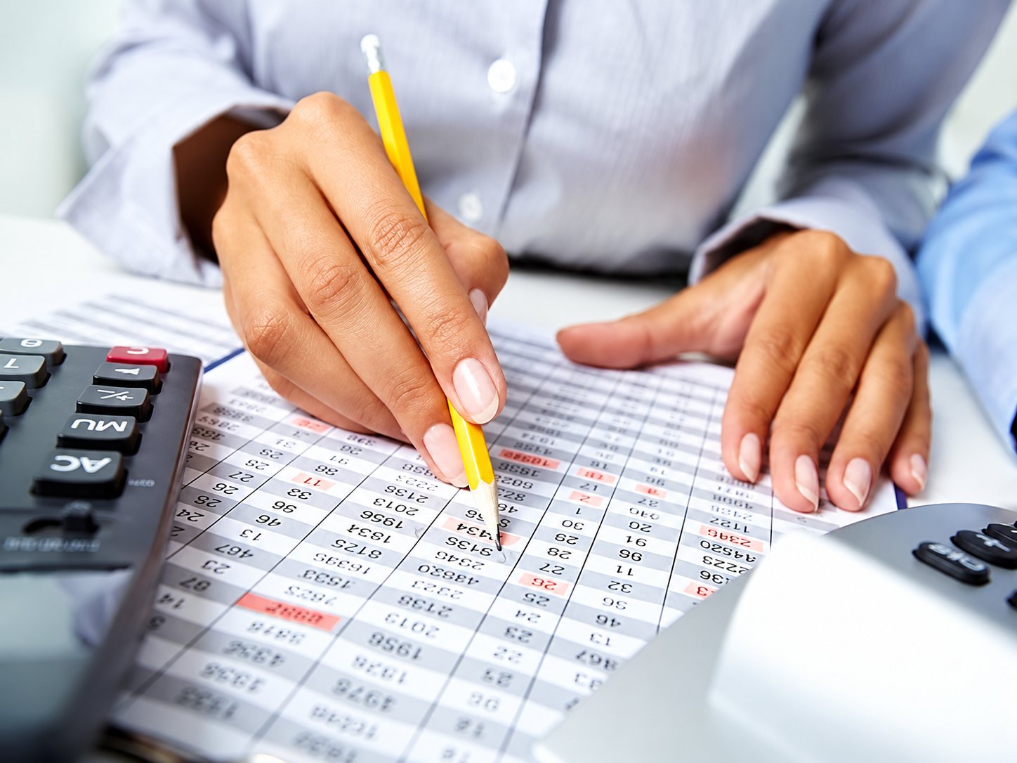 Une personne tient un crayon jaune et suit des lignes de chiffres sur une feuille de calcul imprimée, à côté d'une calculatrice.