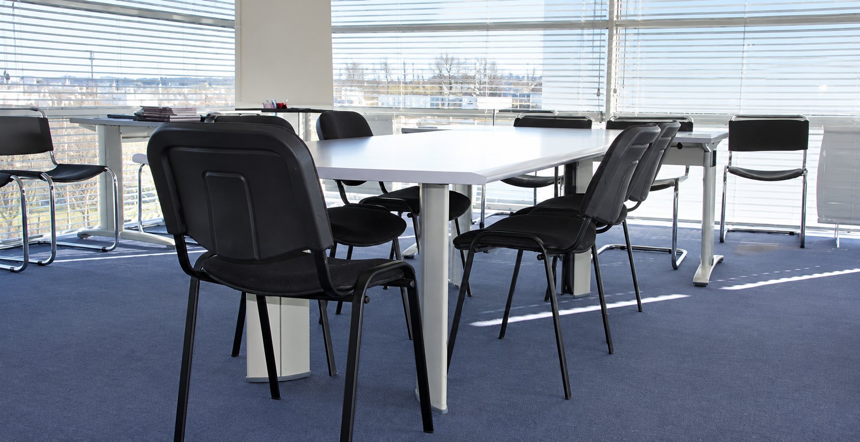 Une salle de réunion lumineuse avec une table blanche entourée de chaises noires et de grandes fenêtres à stores.